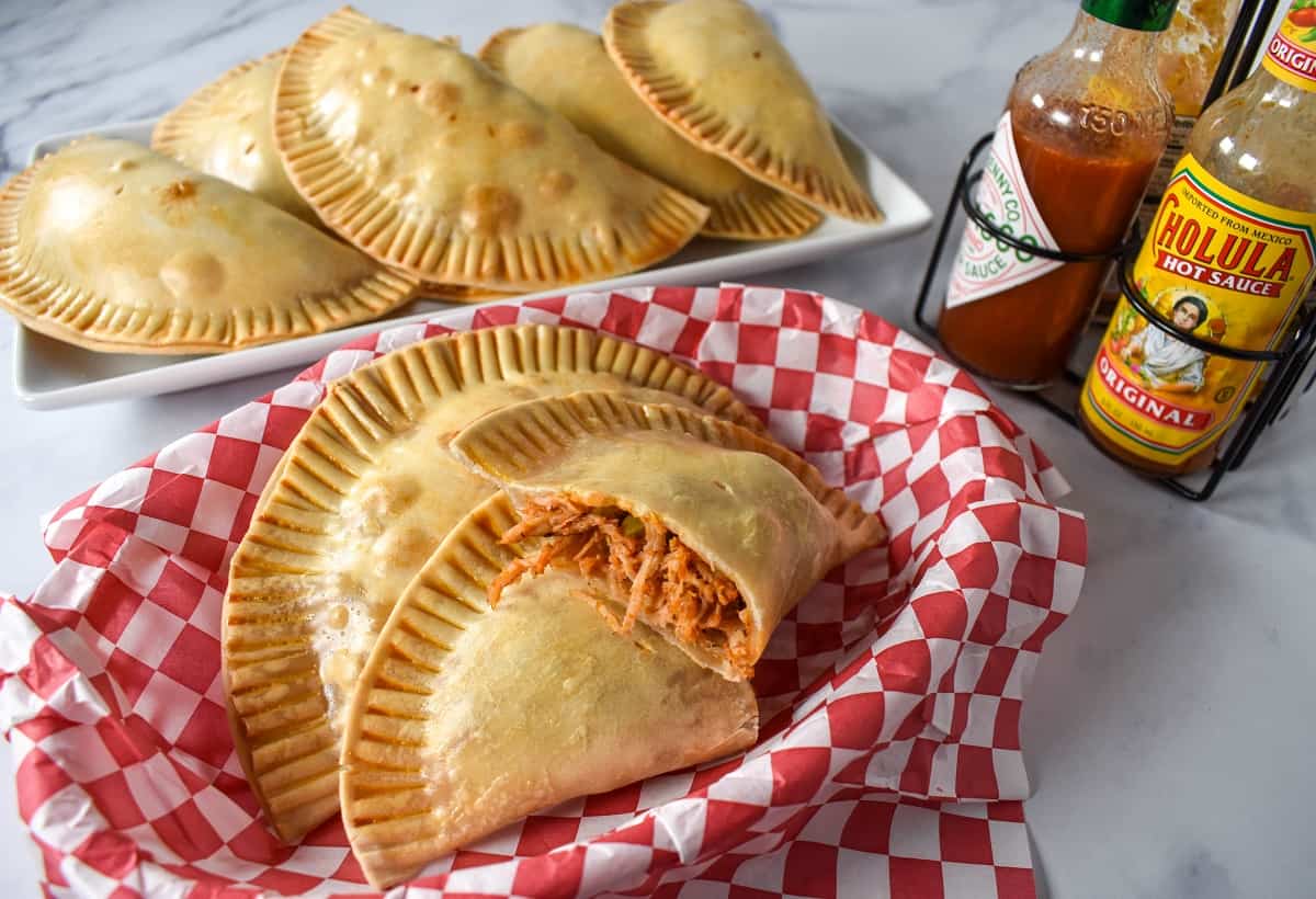 Chicken empanadas in a basket lined with a red and white checkered paper with hot sauce and a platter of empanadas in the background.