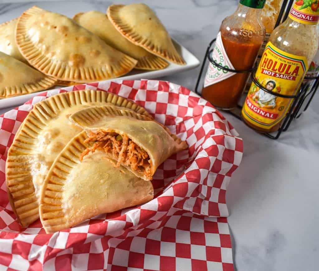 Chicken empanadas served in a basket lined with red and white checkered paper with hot sauces and a platter of empanadas in the background.