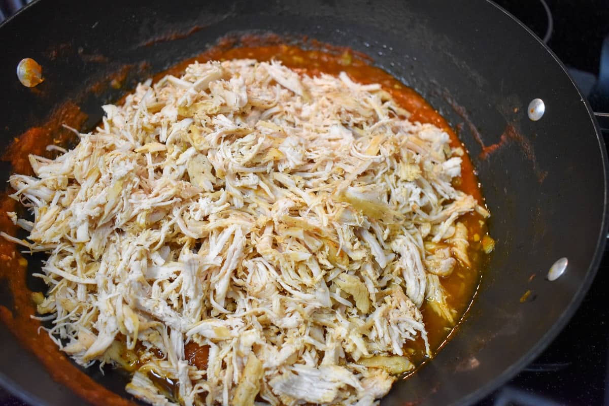 Shredded chicken on tomato sauce before being stirred, in a large, black skillet.