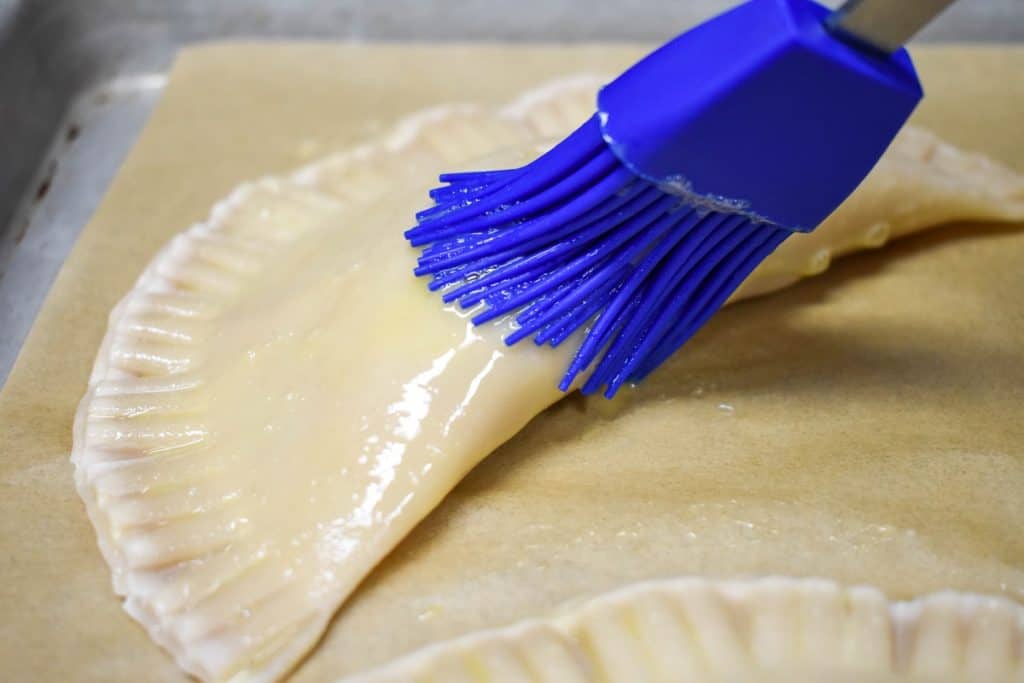 An empanada being coated lightly with egg wash using a blue pastry brush