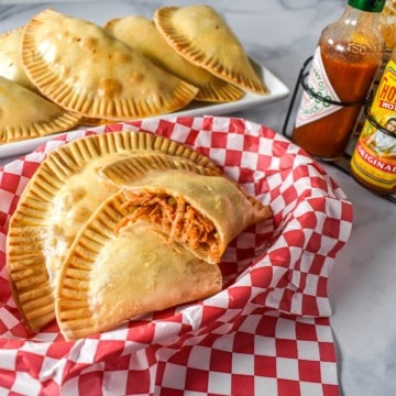 Chicken empanadas in a basket lined with a red and white checkered paper with hot sauce and a platter of empanadas in the background.