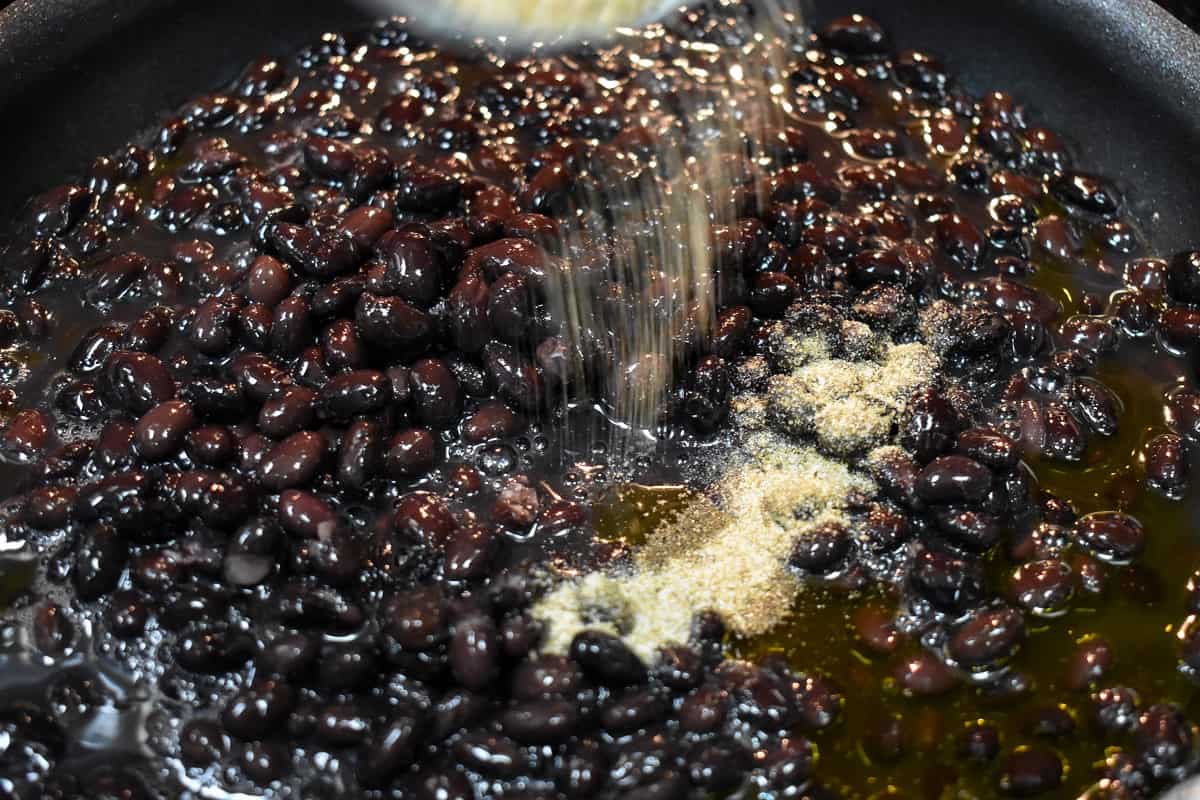 Seasoning being sprinkled on black beans in a black non-stick skillet.