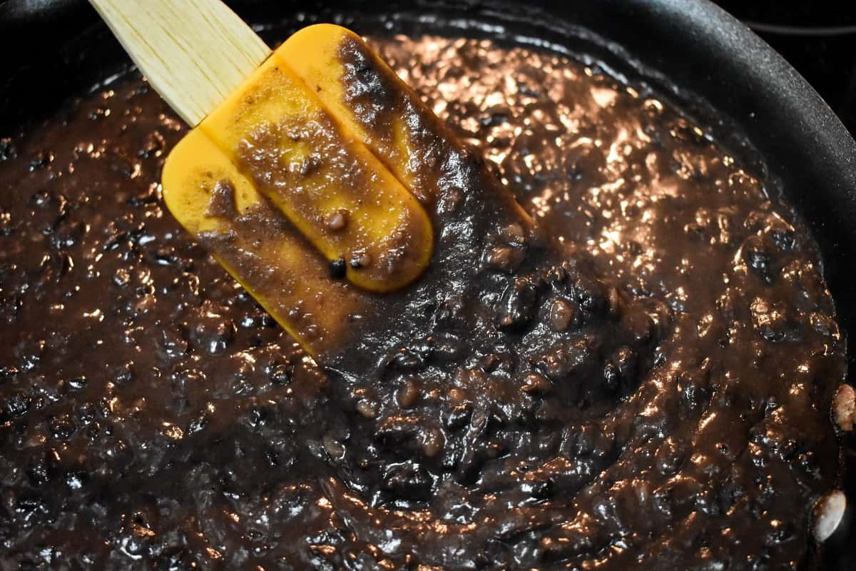 Black beans being mashed in a black non-stick skillet with an orange rubber spatula.