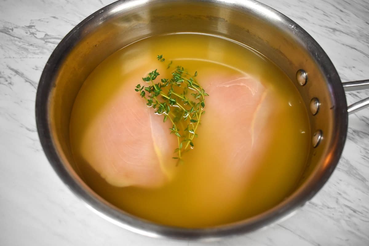 Two raw chicken breasts in a medium saucepan covered with chicken broth and thyme sprigs.