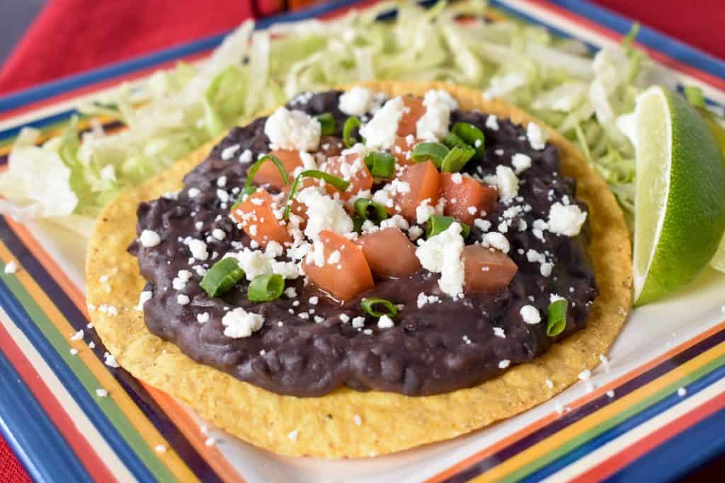 Black refried beans spread on a yellow corn tostada, topped with diced tomatoes, crumbled queso fresco and green onions. The tostada is set on shredded lettuce with a lime wedge on the side.
