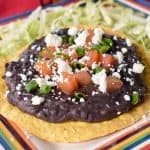 Black refried beans spread on a yellow corn tostada, topped with diced tomatoes, crumbled queso fresco and green onions. The tostada is set on shredded lettuce with a lime wedge on the side.