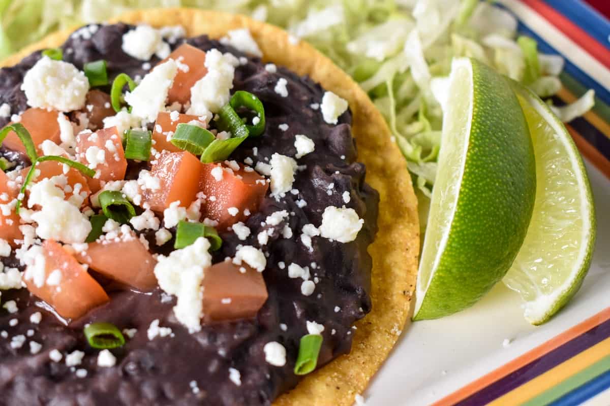 Black refried beans spread on a yellow corn tostada, topped with diced tomatoes, crumbled queso fresco and green onions. The tostada is set on shredded lettuce with two lime wedges on the side.