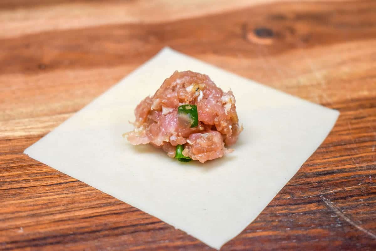 An image of a wonton wrapper with a rounded teaspoon of meat in the center and set on a wood cutting board.