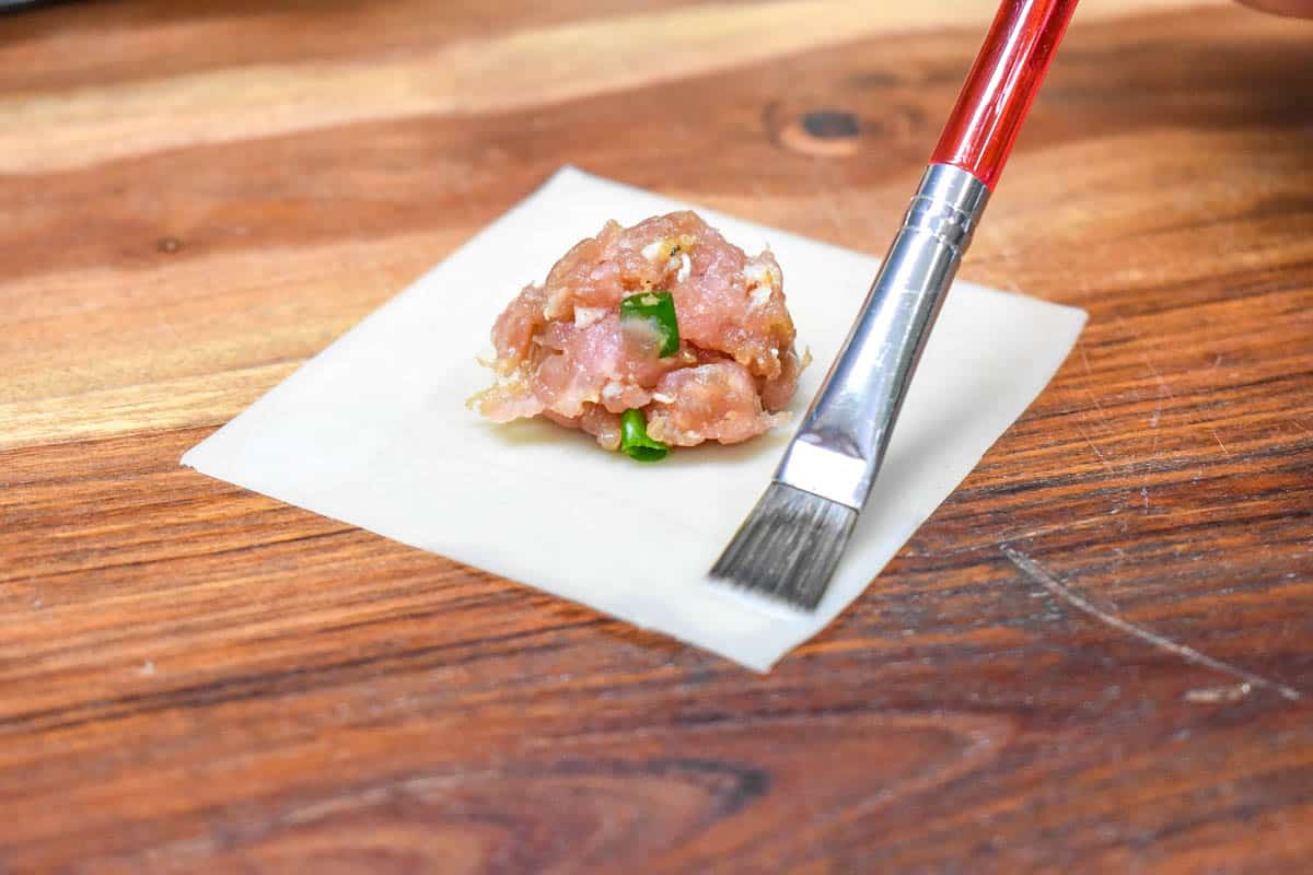 An image of a pastry brush on the edge of a wonton wrapper that has a rounded teaspoon of meat mixture in the center.