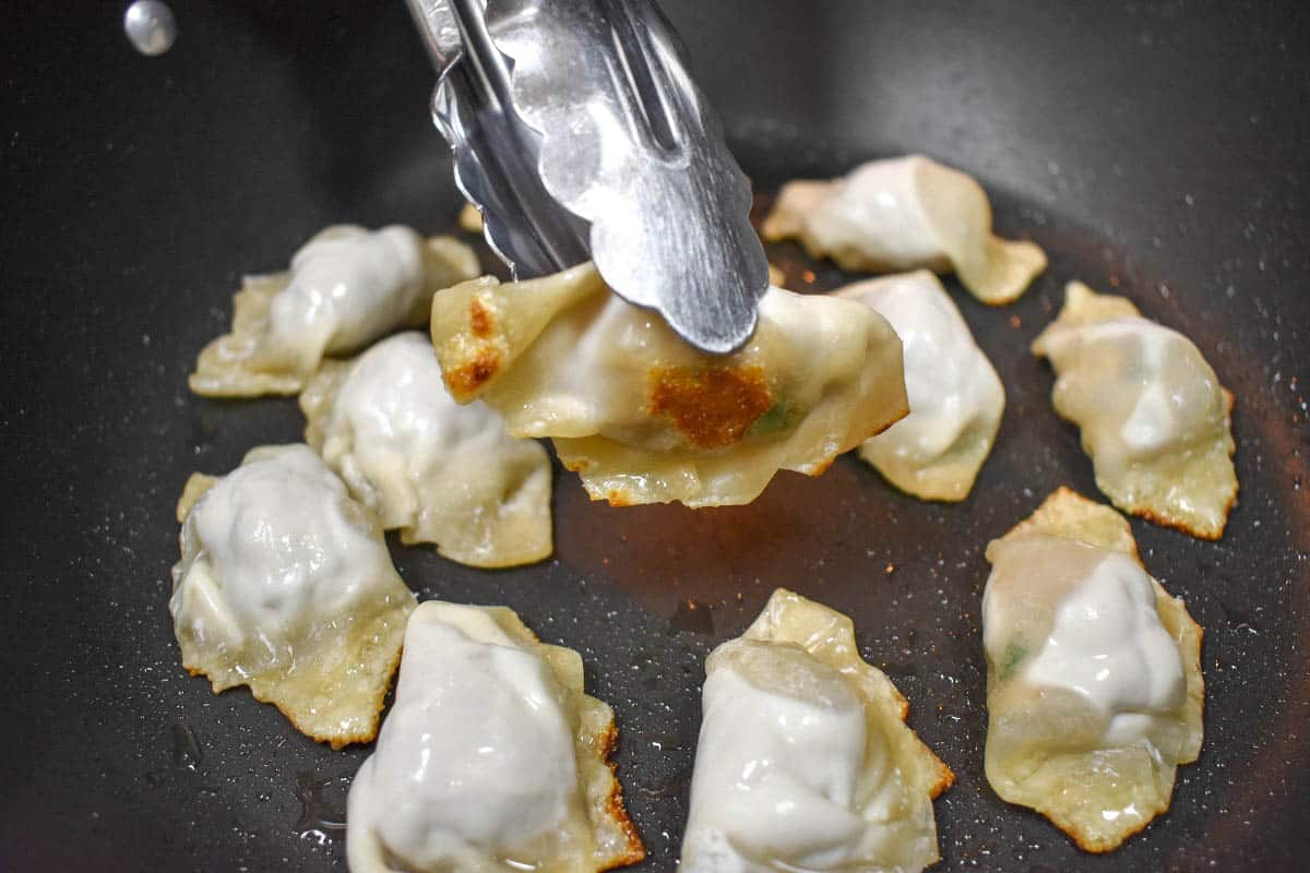Potstickers frying in a large black skillet with one potsticker being lifted with tongs.