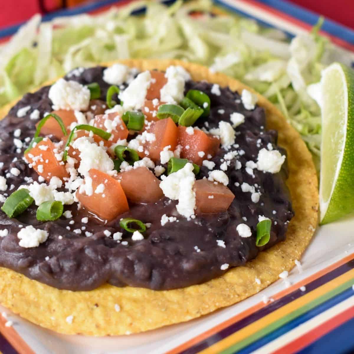 Black refried beans spread on a yellow corn tostada, topped with diced tomatoes, crumbled queso fresco and green onions. The tostada is set on shredded lettuce with a lime wedge on the side.