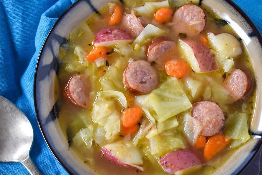 Cabbage sausage soup served in a beige and black bowl set on a blue cloth napkin.