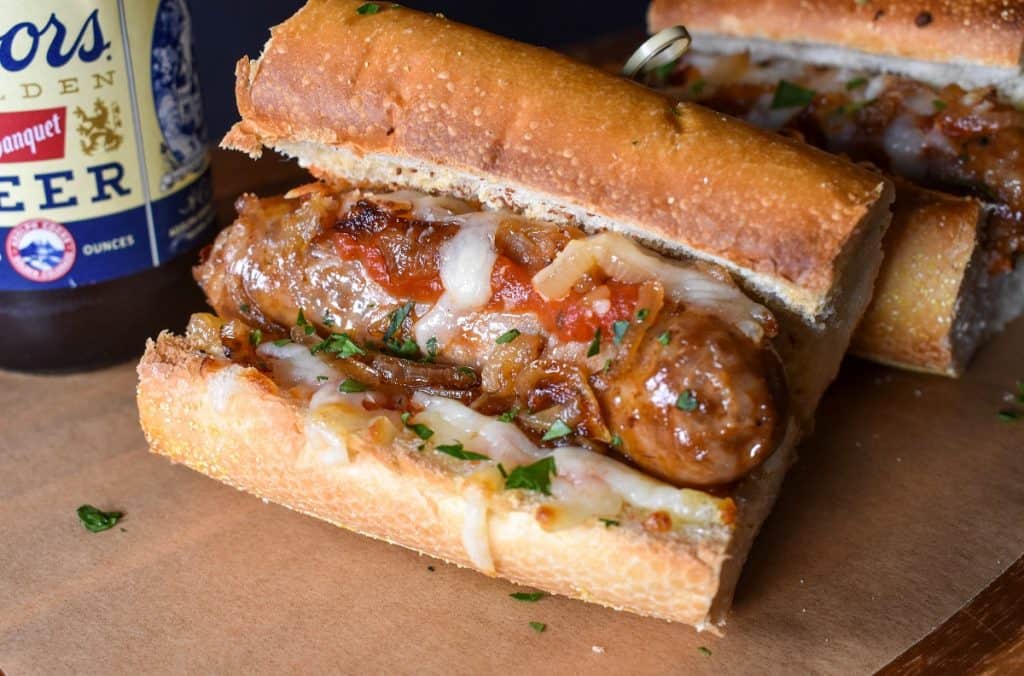 An Italian sausage served on a toasted roll with cooked onions, marinara sauce and shredded mozzarella cheese. It's served on a wood board with beer in the background.