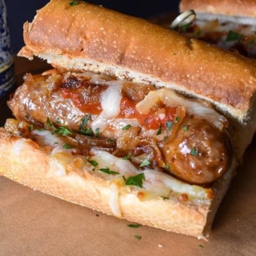 An Italian sausage served on a toasted roll with cooked onions, marinara sauce and shredded mozzarella cheese. It's served on a wood board with beer in the background.