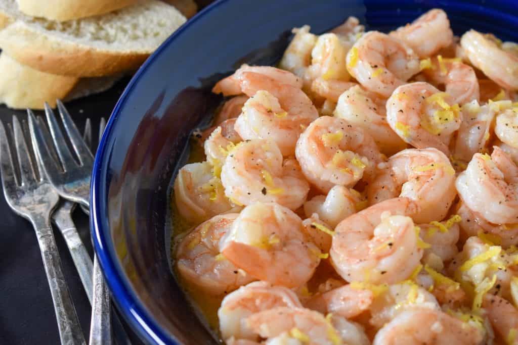 Lemon pepper shrimp served in a blue bowl with small forks on the side and toasted bread rounds in the background.