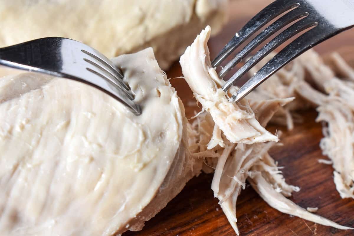 Poached chicken on a cutting board being shredded using two forks.