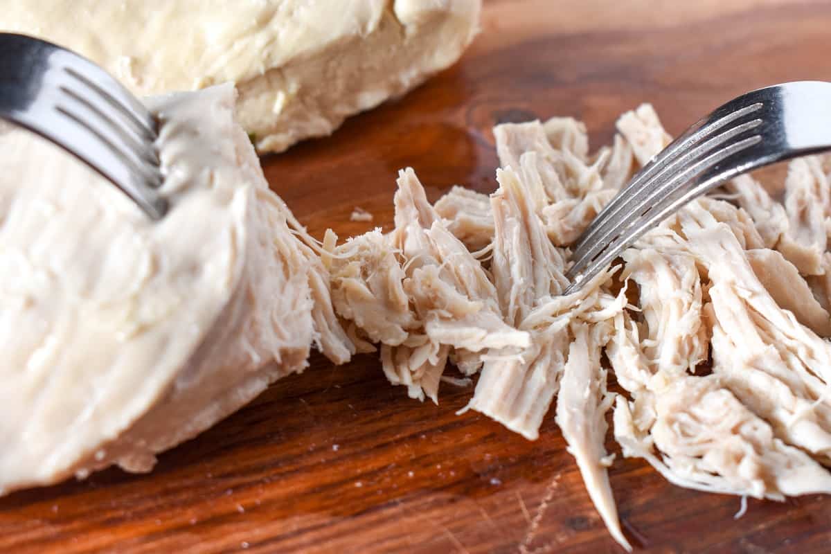 Poached chicken being shredded on a wood cutting board.