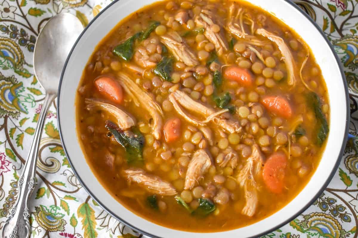 A close up image of lentil soup with shredded chicken served in a white bowl with a spoon to the left had side.
