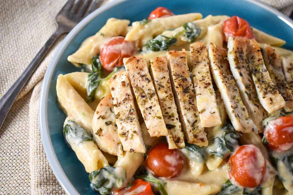 A close up image of creamy penne pasta with greens and grape tomatoes topped with sliced chicken and served in a light blue bowl.