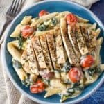 Creamy penne pasta with greens and grape tomatoes topped with sliced chicken cutlets served in a light blue plate.
