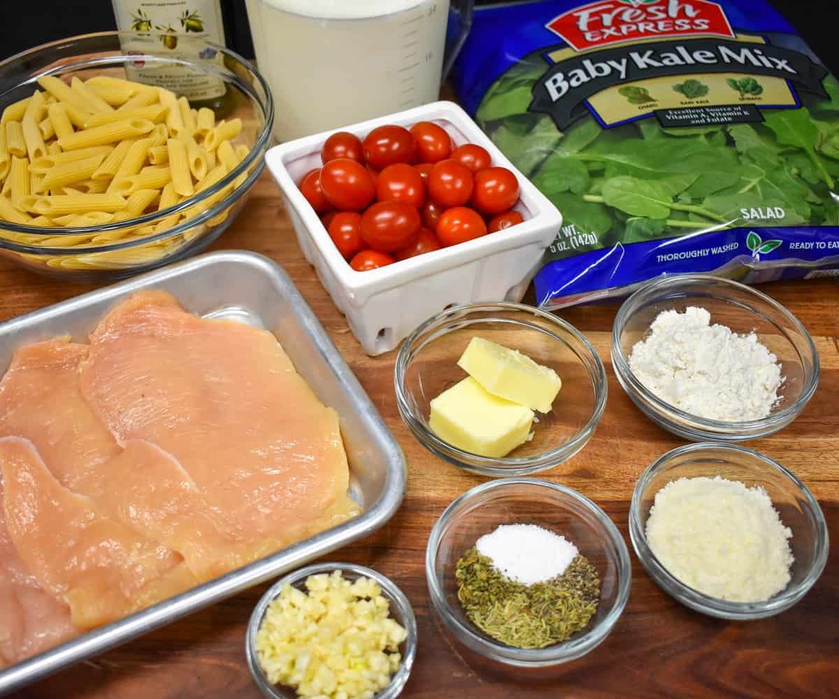 The ingredients for the creamy pasta with chicken arranged in glass bowls and displayed on a wood cutting board.