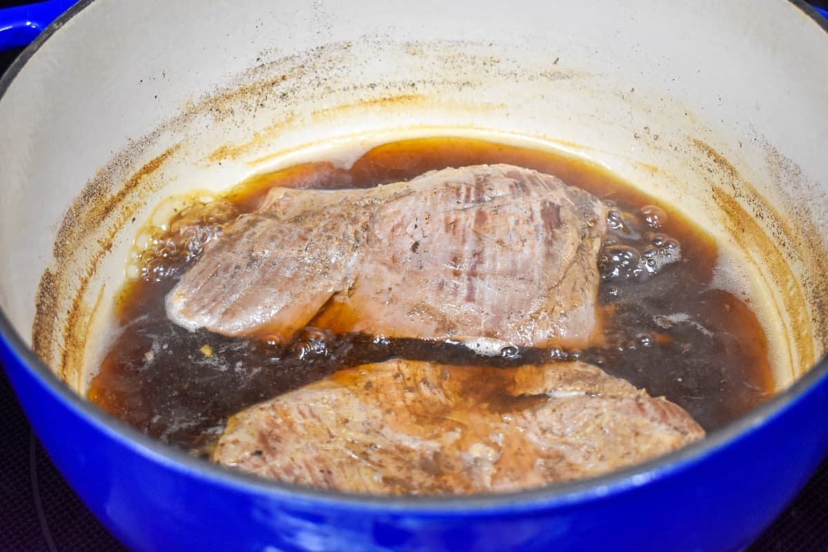 Cooked flank steak in beef broth in a blue and white pot.