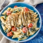 Creamy penne pasta with greens and grape tomatoes topped with sliced chicken cutlets served in a light blue plate.