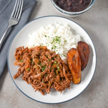 Ropa vieja served with white rice and fried sweet plantains on a white plate with black beans in a small bowl above.