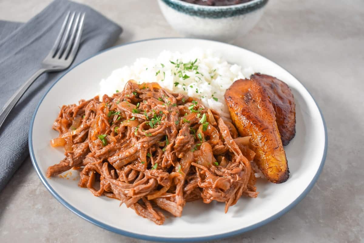 Ropa vieja served with white rice and fried sweet plantains on a white plate.