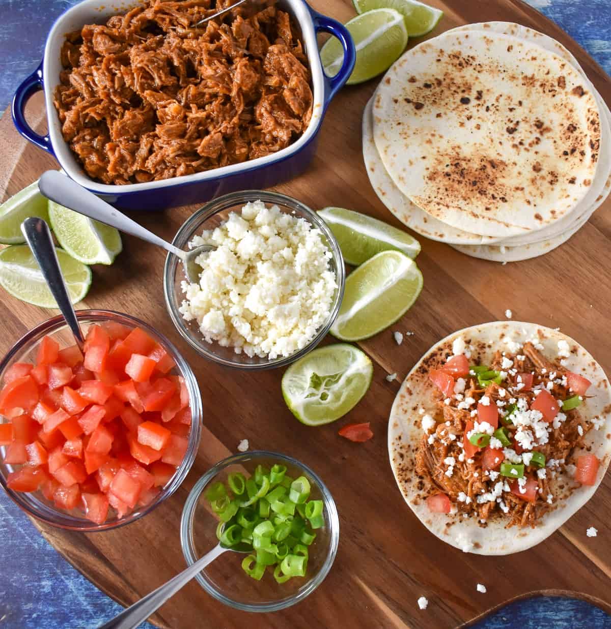 Shredded pork and taco fixings displayed on a wood cutting board.