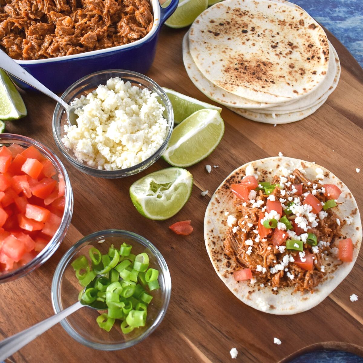 Shredded pork and taco fixings displayed on a wood cutting board.