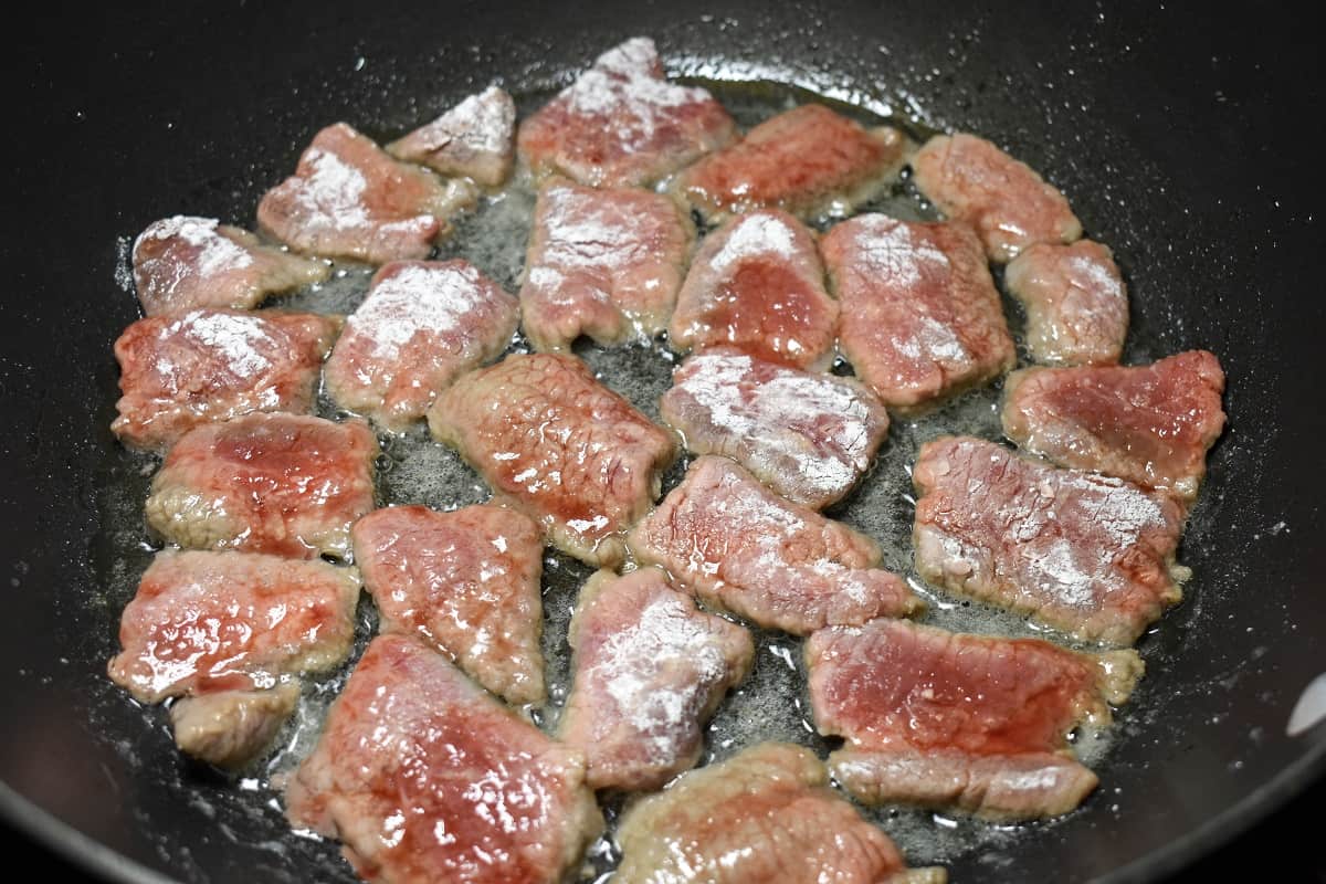 Beef slices that are coated in flour and cornstarch arranged in a black, non-stick skillet.