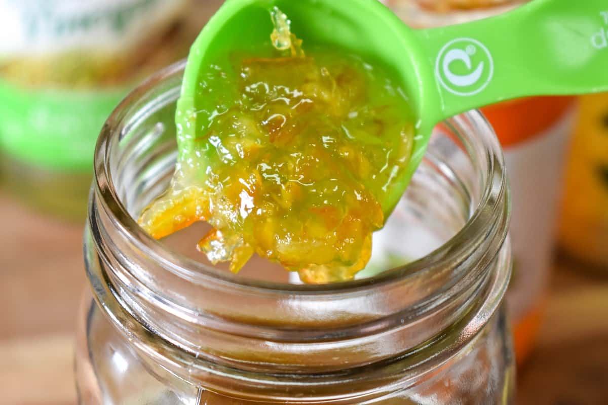 A green tablespoon adding orange marmalade to a glass canning jar.