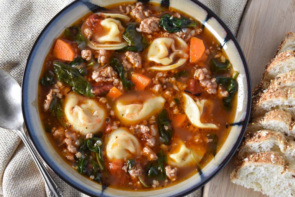 The sausage tortellini soup served in a beige and black rimmed bowl with sliced bread to the right and a spoon to the left on a light colored linen.