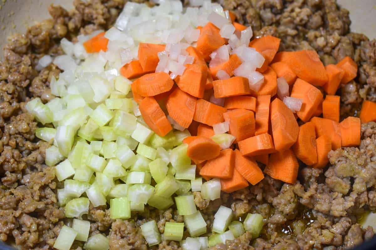 A close up image of browned sausage, diced onions, carrots and celery in a pot.