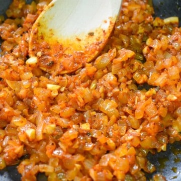 Diced onions, garlic and spices cooked with tomato paste in a black non-stick skillet with a wooden spoon in the skillet.