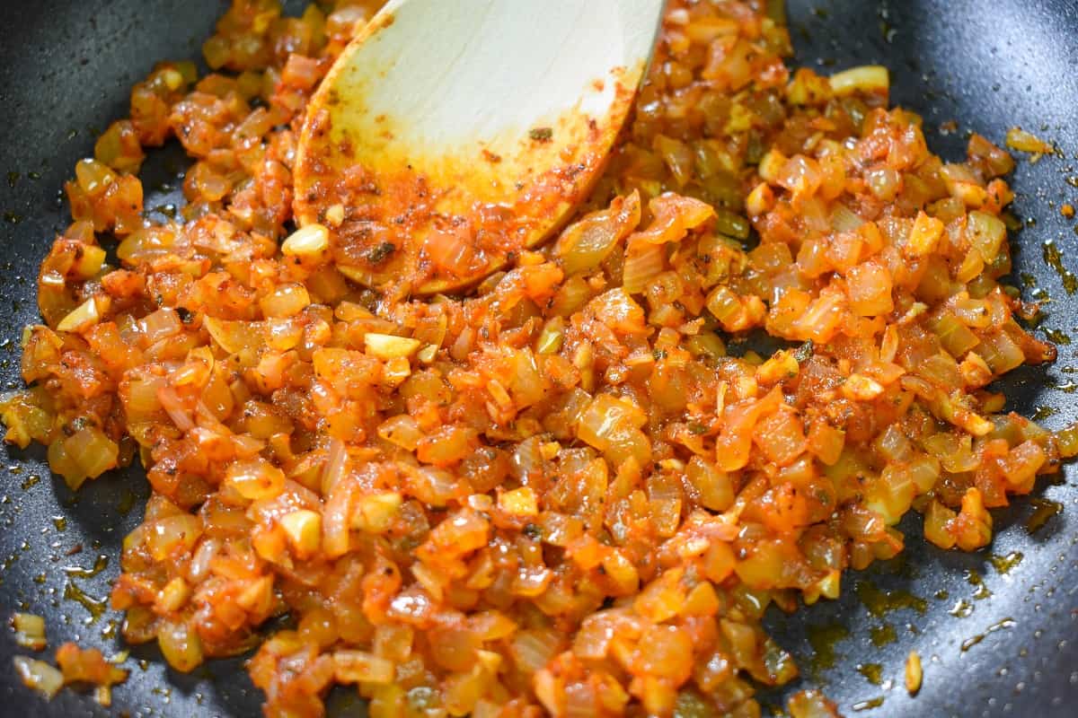 Diced onions, garlic and spices cooked with tomato paste in a black non-stick skillet with a wooden spoon in the skillet.