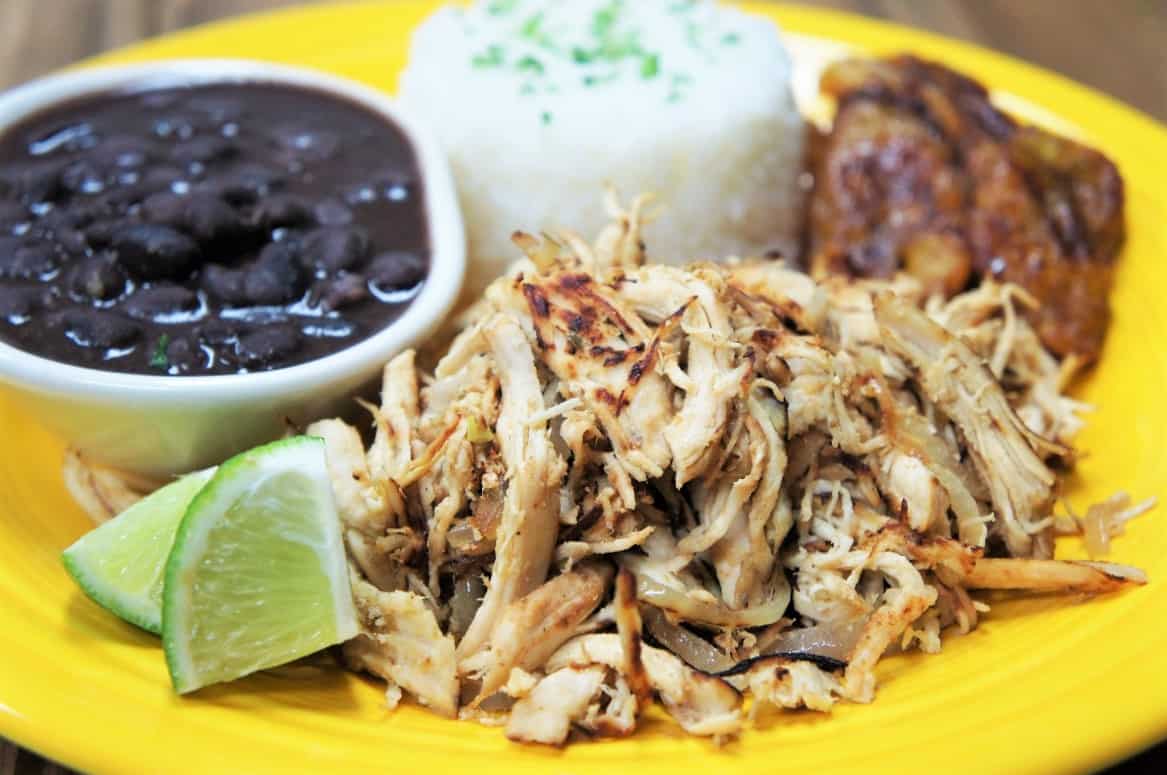 Shredded chicken served with white rice, black beans, plantains and lime wedges on a yellow plate.