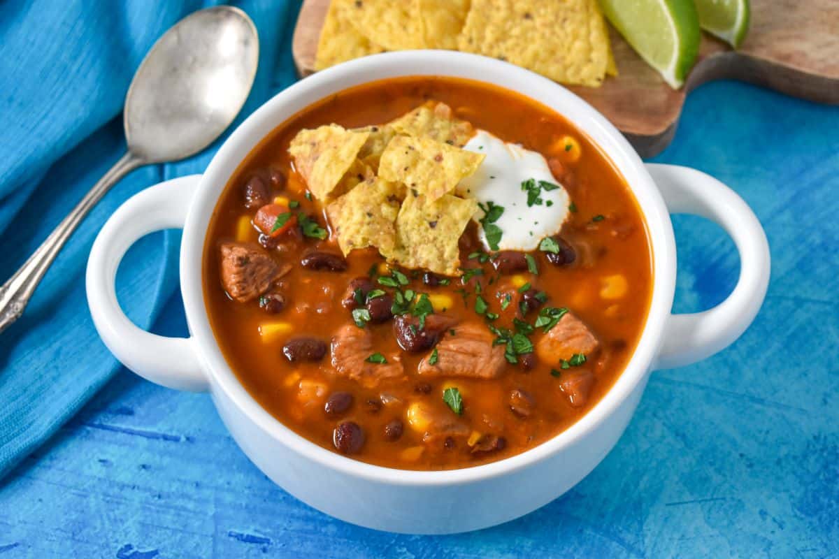 The soup served in a white bowl garnished with parsley, sour cream, and tortilla chips with extra chips, lime wedges and a spoon in the background.