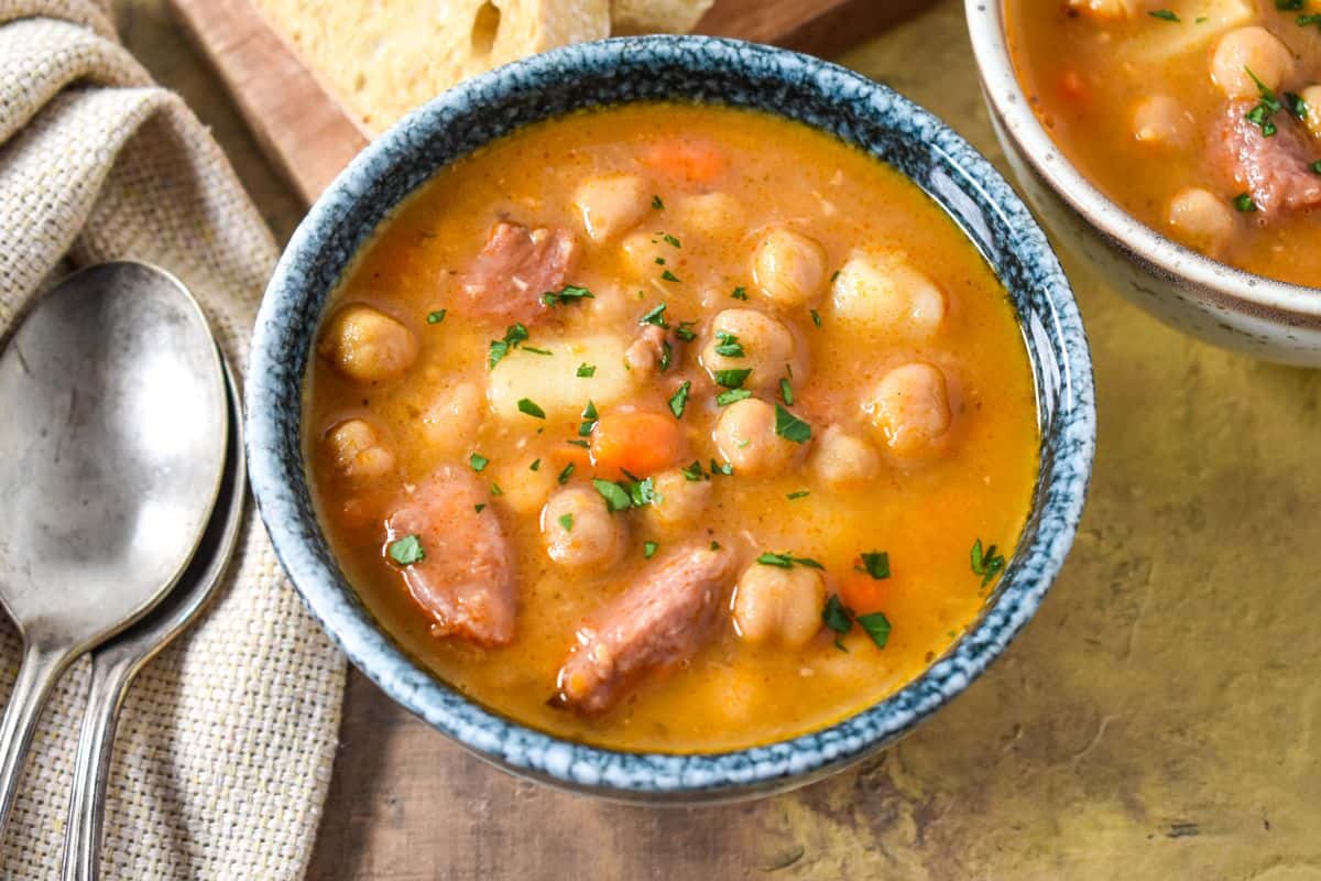 Close-up of a bowl of soup with chickpeas, potatoes, and smoked pork in broth.