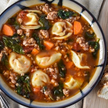 The sausage tortellini soup served in a beige and black rimmed bowl with sliced bread to the right.