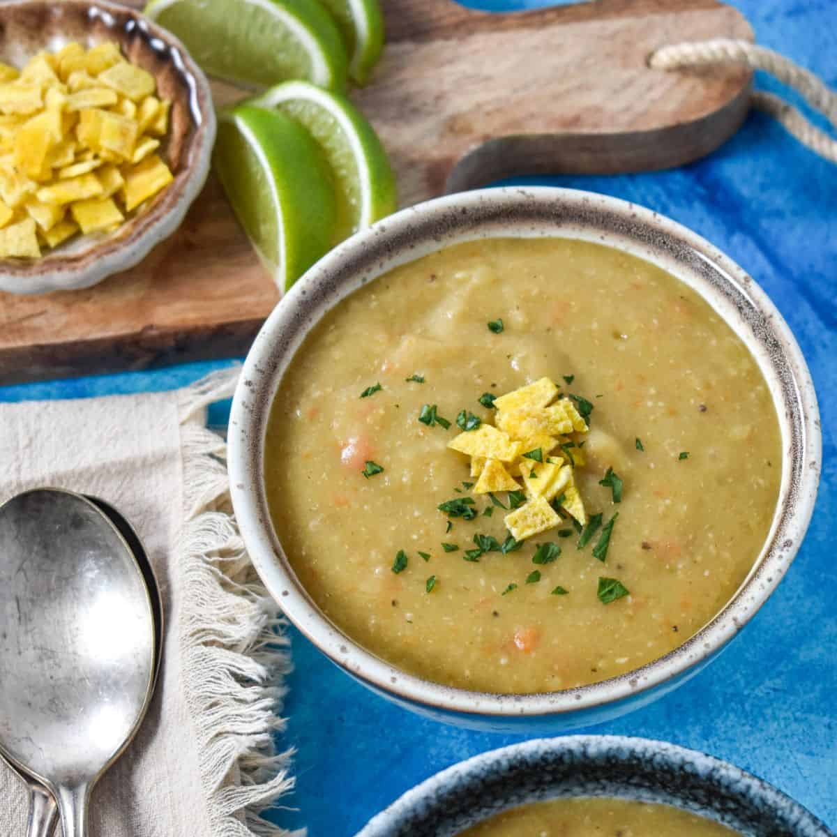 A bowl of thick, creamy soup topped with crushed plantain chips and fresh herbs, served with lime wedges on a wooden board.