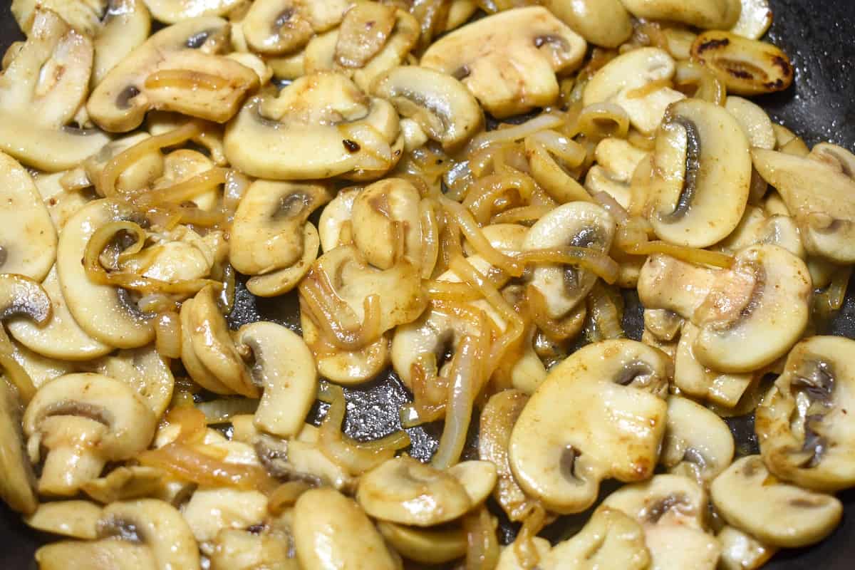 A close up of sliced onions and mushrooms cooking in a black skillet. Both the mushrooms and onions are a golden color already.