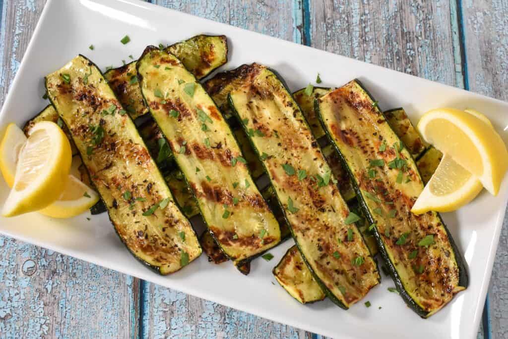 Sliced grilled zucchini arranged on a large white platter, garnished with chopped parsley and lemon wedges on the corners.