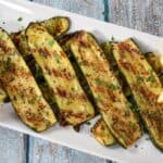 Sliced grilled zucchini arranged on a large white platter, garnished with chopped parsley and lemon wedges on the corners.