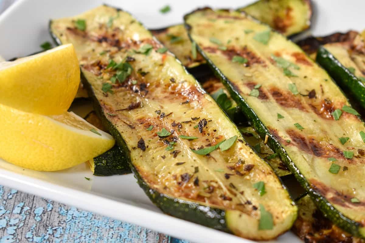 A close up image of sliced grilled zucchini arranged on a white platter, garnished with chopped parsley and lemon wedges on the corner.