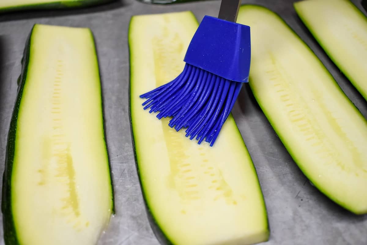 Thick zucchini slices on a metal baking sheet being brushed with olive oil with a blue silicone pastry brush.