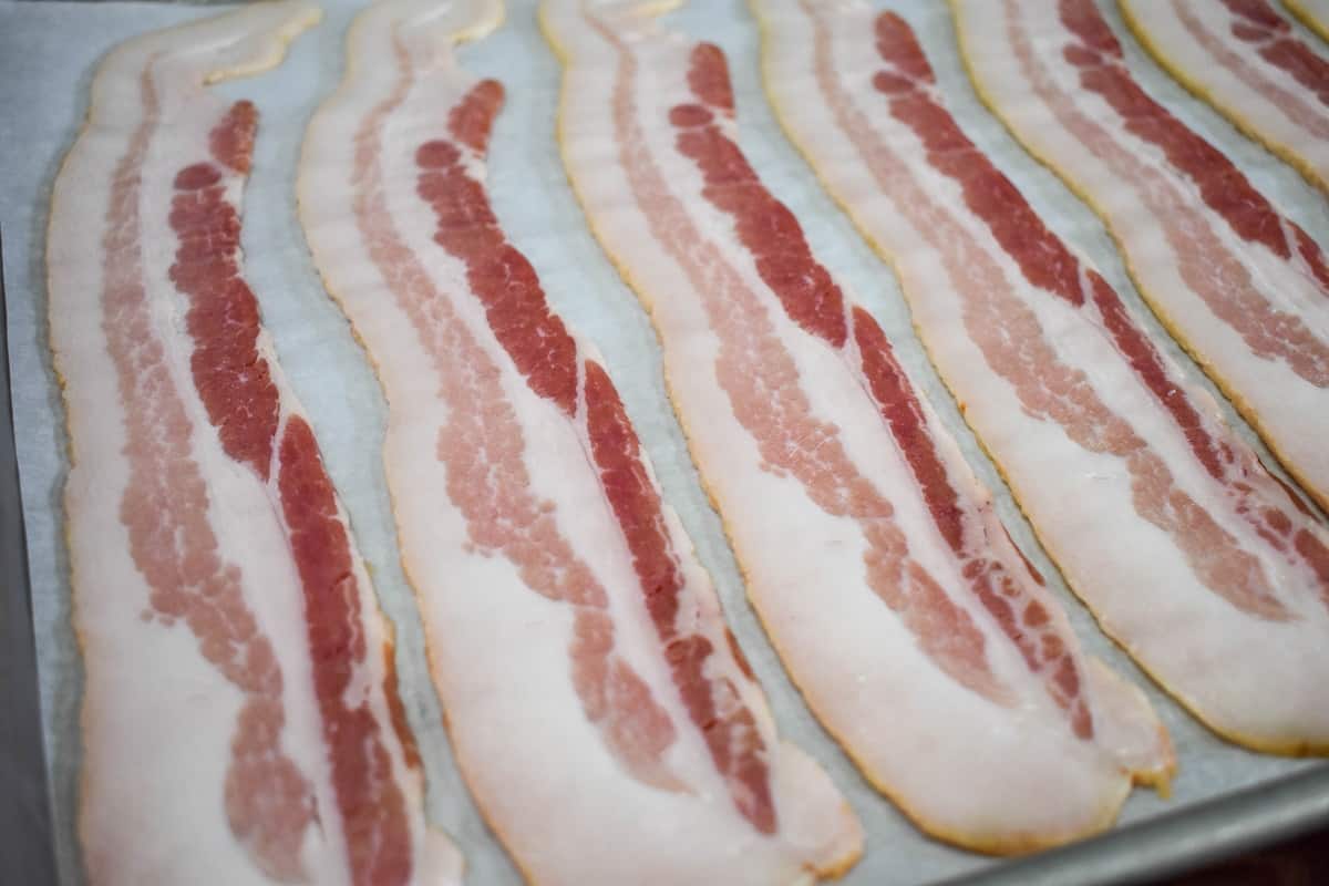 Uncooked bacon slices arranged on a large baking sheet that's lined with white parchment paper.