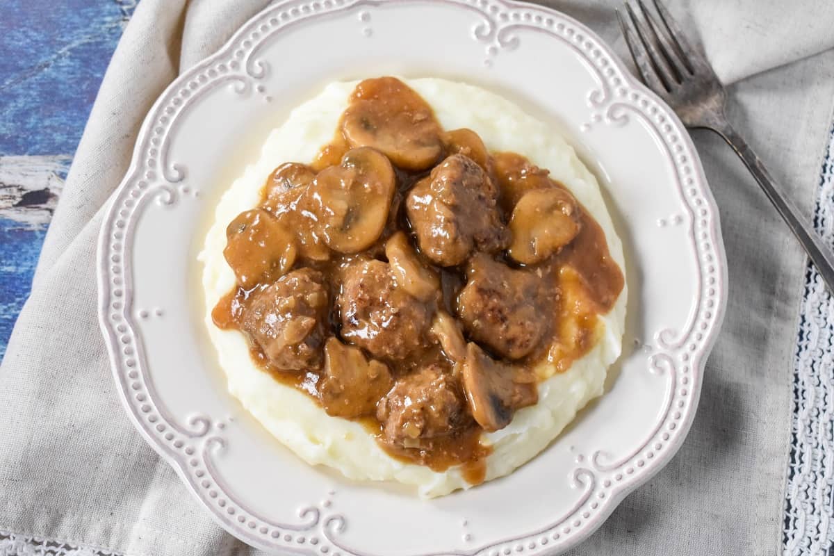 Salisbury steak meatballs and mushroom gravy on a bed of mashed potatoes served on a white plate with an off-white linen and a fork to the right side.