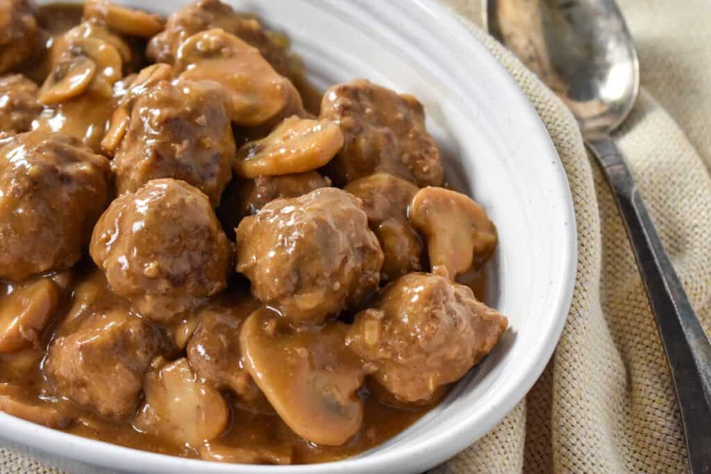 Meatballs and a mushroom gravy served in an oval white bowl on a cream colored linen with a serving spoon on the right side.